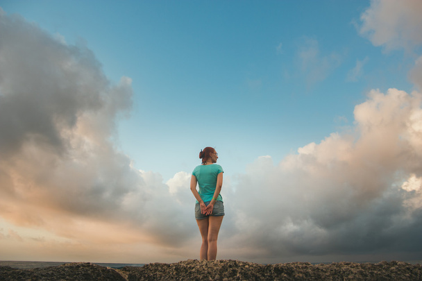 Jovem mulher em shorts e camiseta azul em pé na costa olhando para um mar, as nuvens se parecem com asas
 - Foto, Imagem