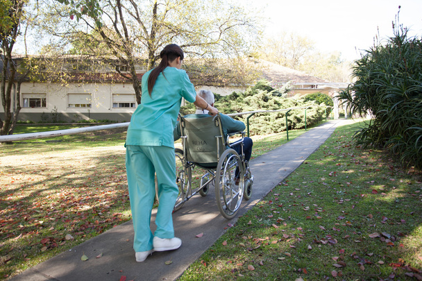 Krankenschwester trägt eine Patientin im Rollstuhl im Park - Foto, Bild