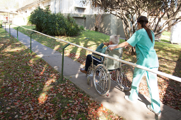 Krankenschwester trägt eine Patientin im Rollstuhl im Park - Foto, Bild
