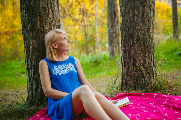 blonde girl in a blue dress sitting on a red blanket on top of the green grass, reading a book in the middle of pine forest trees - Zdjęcie, obraz