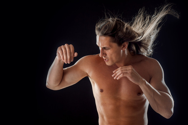 muscular man with long hair, clasps hands in fist, black background - Photo, Image