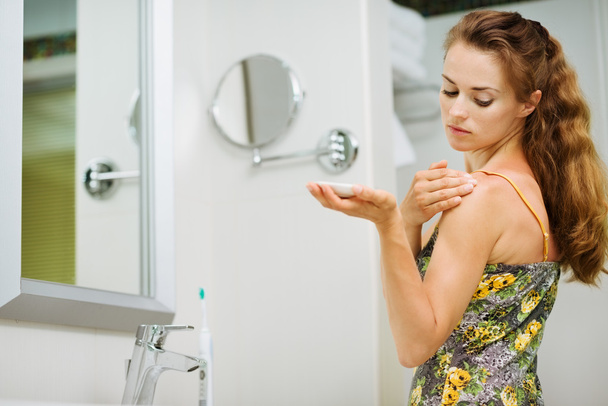 Молодая женщина наносит крем для тела на плечо в ванной комнате
 - Фото, изображение