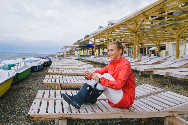 young woman in a red raincoat and blue rubber boots sitting on a wooden deck chair by the stormy  - Foto, afbeelding