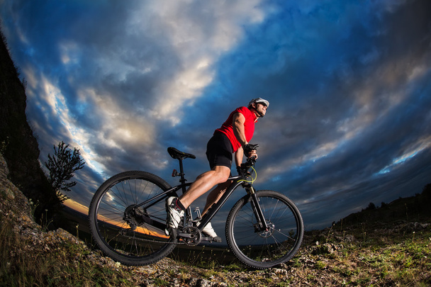 pyöräilijä seisoo maastopyörä polulla auringonlaskun aikaan
 - Valokuva, kuva