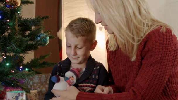 Happy son with mother sitting under new year tree and play with snowman - Záběry, video