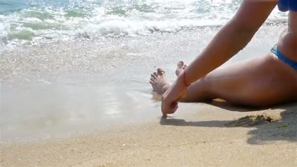 Mujer dibujando corazón en la arena de la playa, olas salpicando en su cuerpo, piernas solamente
 - Metraje, vídeo