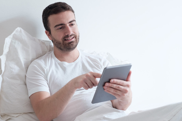Ο άνθρωπος χρησιμοποιώντας τον ηλεκτρονικό tablet ξαπλωμένη στο κρεβάτι - Φωτογραφία, εικόνα