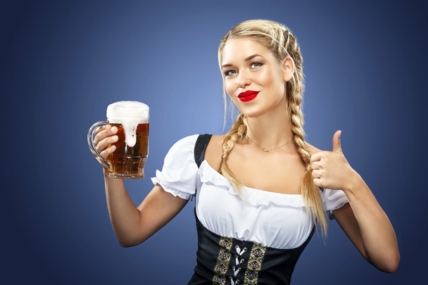 Young sexy Oktoberfest waitress, wearing a traditional Bavarian dress, serving big beer mugs on blue background. - Фото, изображение