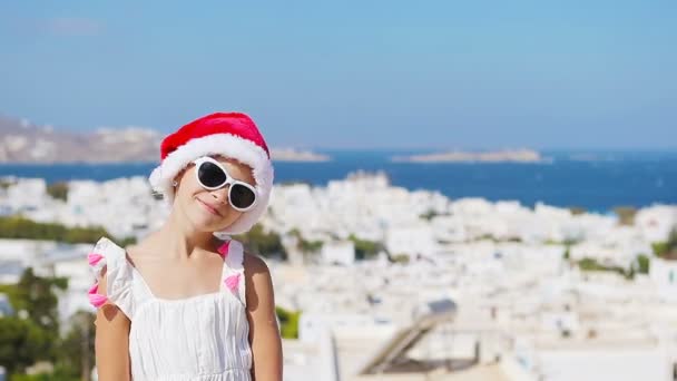 Pikkutyttö jouluhattu päässä ulkona taustalla vanha katu Mykonos. Lapsi tyypillisen kreikkalaisen perinteisen kylän kadulla, jossa valkoiset seinät ja värikkäät ovet Mykonoksen saarella, Kreikassa
 - Materiaali, video