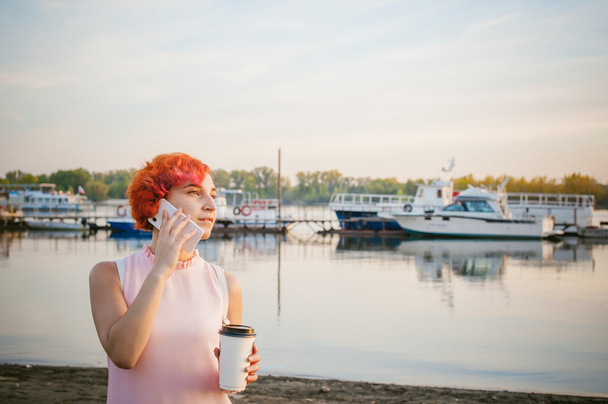 girl in pale pink dress with red hair and backpack walking along river bank, talking on the phone and drinking coffee from a cardboard cup, against backdrop of boats moored on a warm summer day - Fotoğraf, Görsel