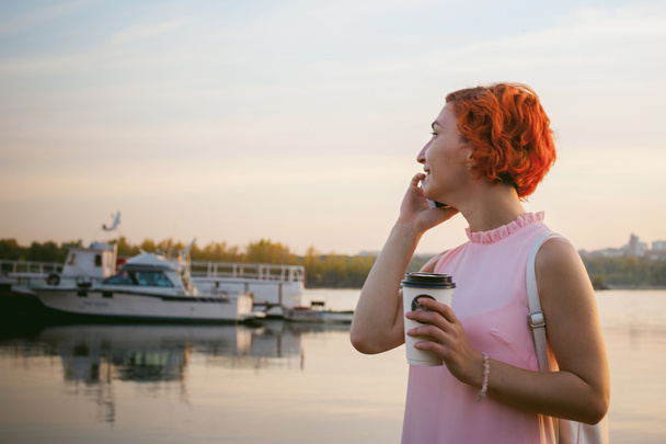 girl in pale pink dress with red hair and backpack walking along river bank, talking on the phone and drinking coffee from a cardboard cup, against backdrop of boats moored on a warm summer day - Fotoğraf, Görsel