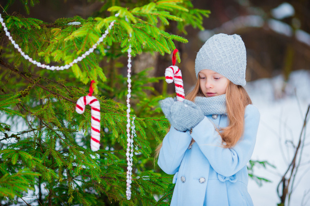 Portrait of a little cute smiling girl in a blue coat and mittens standing near the decorated Christmas tree on a frosty winter day outdoor. - Photo, Image