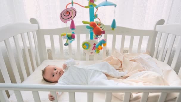 Lindo bebé niño acostado en una cama de madera blanca y jugando con un juguete móvil
 - Imágenes, Vídeo