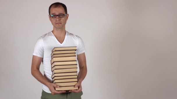 Nerdy man in black rim glasses carrying big stack of books against gray background, isolated. 4K shot - Video