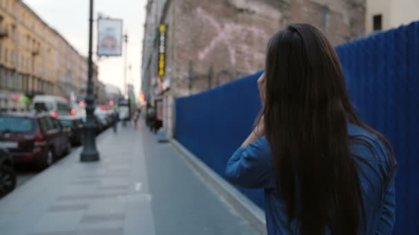 Lady walks in the street near blue fence. Backview of woman with long hair talking on the phone. Slow mo, steadicam shot - Materiał filmowy, wideo