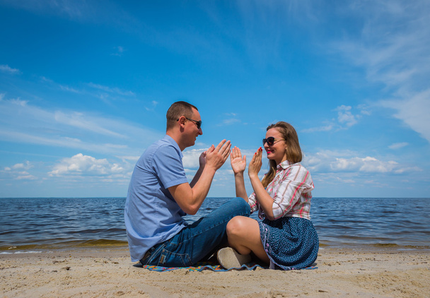 a loving couple sittingon the beach, near the water, playing with each other, blue sky on the background, sunny day, wide angle, long white clouds - Photo, Image