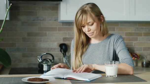 Young attractive woman reading a book in the kitchen. Nearby stands a glass of milk. Concept - healthy eating and a healthy lifestyle - Filmati, video