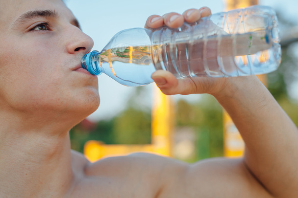 Ο άνθρωπος είναι πόσιμο νερό μετά την προπόνηση. Εξωτερική γυμναστήριο φόντο - Φωτογραφία, εικόνα