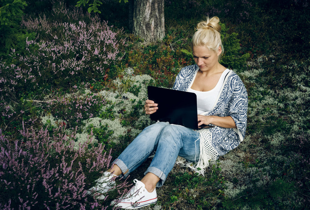森の草の上に座って、コンピュータを探している美しい若い女性。屋外でラップトップを使用している女性. - 写真・画像