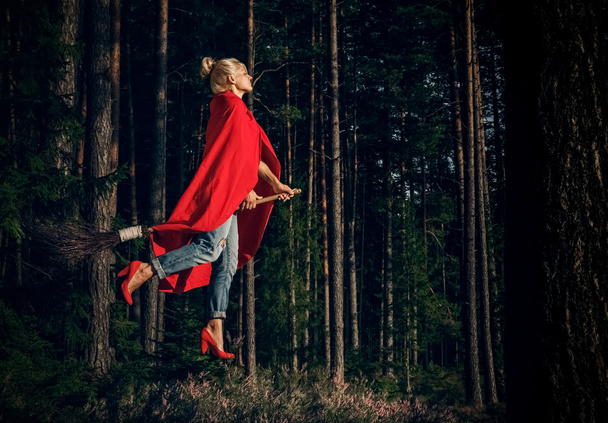 Modern witch blond in jeans and red cape flying on a broom through the forest. Levitation. Halloween holiday. - Foto, Imagen