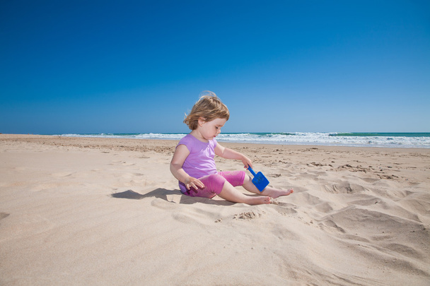 μικρό κορίτσι συνεδρίαση παίζοντας με μπλε φτυάρι στην αμμουδιά - Φωτογραφία, εικόνα