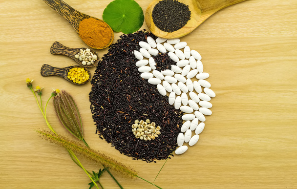 Černá rýže a bílé pilulky tvořící yin yang symbol a bylinné lázně komprese míč, kurkuma prášek, proso, sóji, bazalka semen v vařečku na hnědé dřevěné prolnutí herb a lékařství - Fotografie, Obrázek