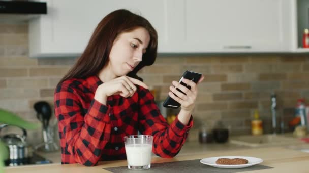 A young woman uses a smartphone. He is sitting in his kitchen, standing next to a glass of milk. Healthy lifestyle - Felvétel, videó