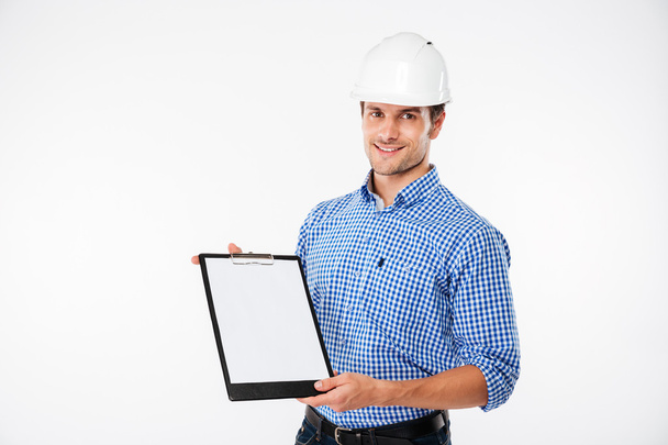 Cheerful young man builder in building helmet holding blank clipboard - Photo, image