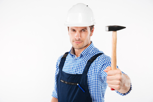 Happy young builder in helmet standing and holding hammer - 写真・画像