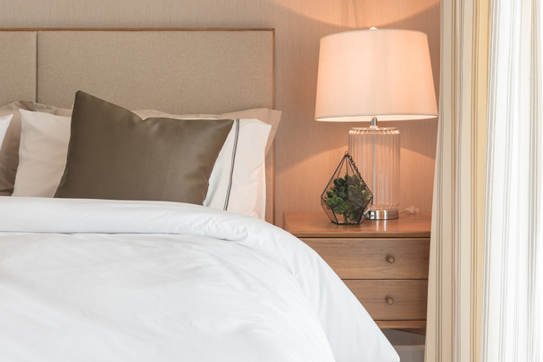 braunes Kissen auf weißem, modernem Bett mit Lampe auf der Tischseite  - Foto, Bild