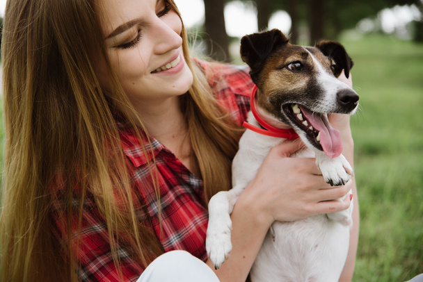 young pretty girl in red tartan shirt and jeans sit at the lawn and hold her dog. - Foto, Bild