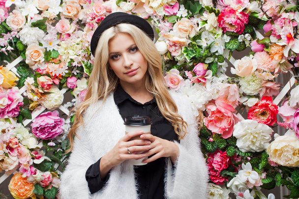 Young beautiful blonde woman in fashionable hat on floral background with coffee to go - Φωτογραφία, εικόνα