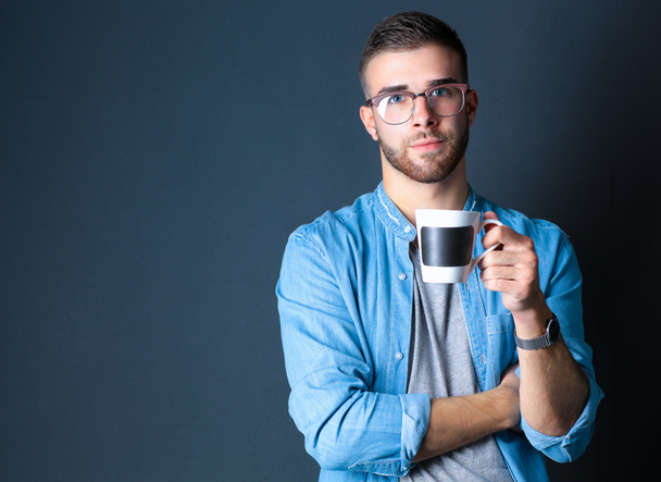 Portrait of a handsome young man standing and holding a cup of coffee in his hands - 写真・画像