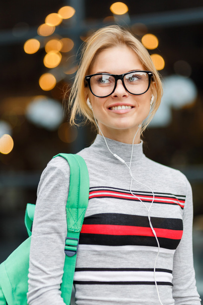 Smiling girl with headphones and backpacker against of glass door - Fotografie, Obrázek