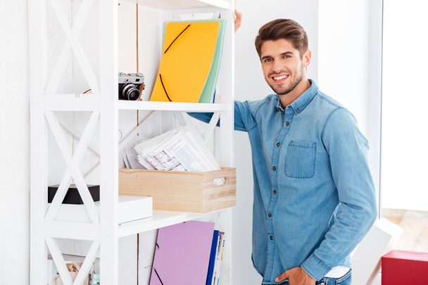 Smiling young man standing at shelf with folders and documents - Foto, Bild