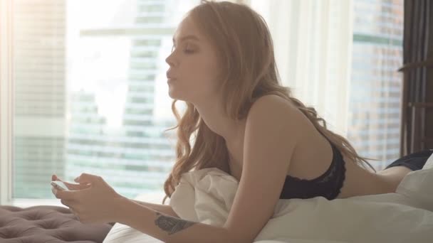Young woman in black lingerie lying in bed with smartphone and sending sms - Πλάνα, βίντεο