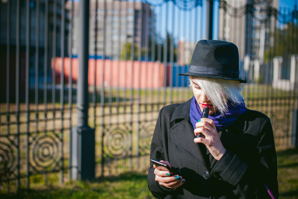 portrait of young beautiful woman with white hair, in a black coat, a skirt and a black hat, smoking an electronic cigarette, blowing the smoke vapor - Photo, image