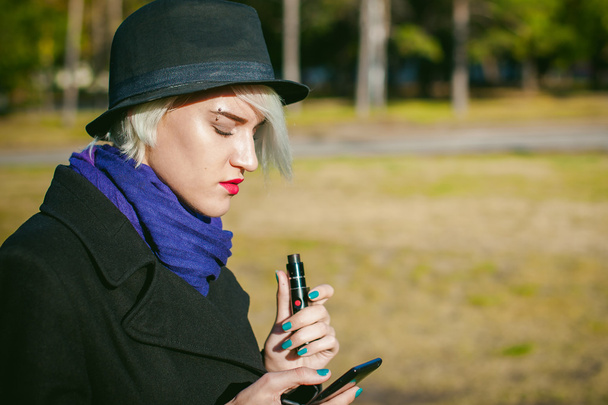 portrait of young beautiful woman with white hair, in a black coat, a skirt and a black hat, smoking an electronic cigarette, blowing the smoke vapor - Фото, изображение