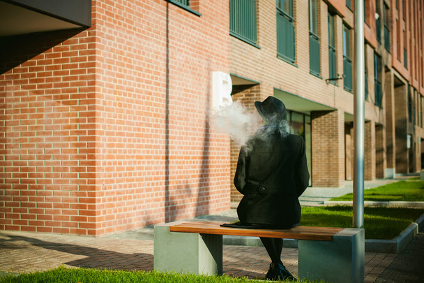 portrait of young beautiful woman with white hair, in a black coat, a skirt and a black hat, smoking an electronic cigarette, blowing the smoke vapor - Valokuva, kuva