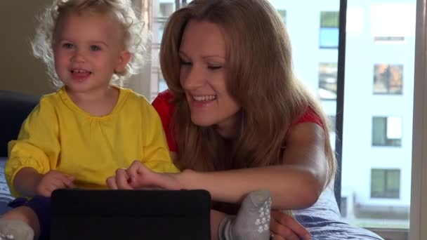 Cute toddler girl and her mom smile using tablet computer lying on sofa - Imágenes, Vídeo