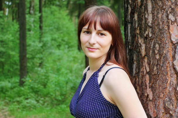 Pretty woman poses near tree in green summer forest at sunny day - Фото, изображение