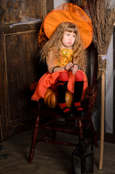 smutna dziewczyna czarownica halloween z długimi włosami w strój i kapelusz w kolorze pomarańczowym, siedząc na dyni na krześle retro z miotłą w stare drewniane drzwi wewnątrz - Zdjęcie, obraz