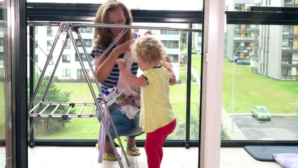 Mother with her toddler daughter hanging washed laundry on stand in balcony - Footage, Video