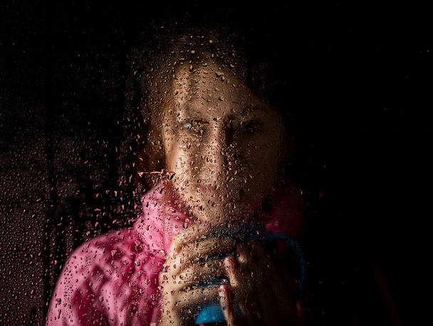 young sad woman portrait behind the window in the rain with rain drops on it. girl holding a cup of hot drink - Photo, image