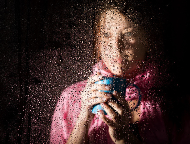 young sad woman portrait behind the window in the rain with rain drops on it. girl holding a cup of hot drink - Foto, Bild