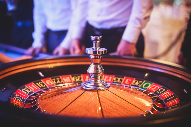 A close-up vibrant image of multicolored casino table with roulette in motion, with the hand of croupier, and a group of gambling rich wealthy people  - Φωτογραφία, εικόνα