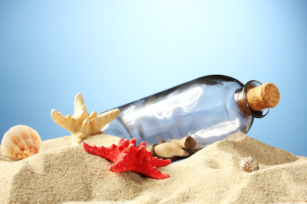 Glass bottle with note inside on sand, on blue background - Photo, Image