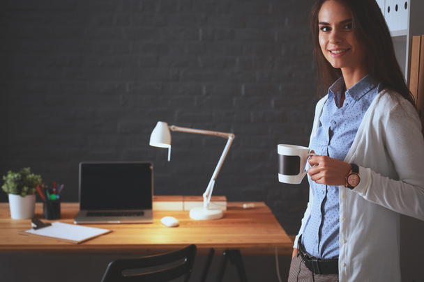 Young woman standing near desk with laptop holding cup of coffee - Fotoğraf, Görsel