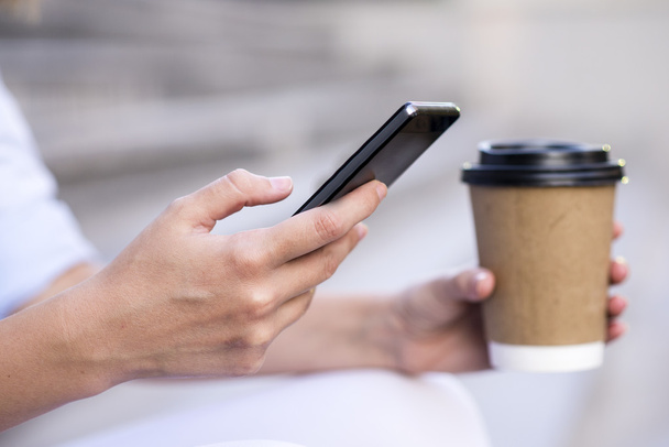 Close up of a businesswoman using mobile phone and holding paper cup. Close-up detail of a businesswoman hand holding paper cup and using a smartphone - Fotó, kép