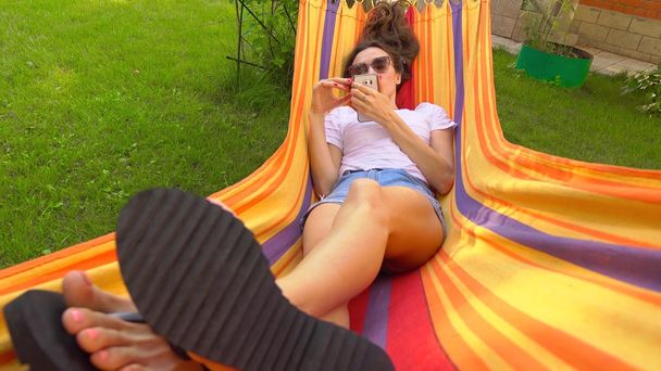 Beautiful caucasian brunette girl swinging in bright hammock and using her smartphone - Valokuva, kuva
